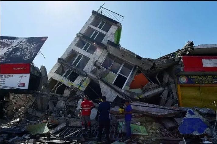 A multi-storey building leaning and broken open above commercial shopfronts, with rubble across the pavement.