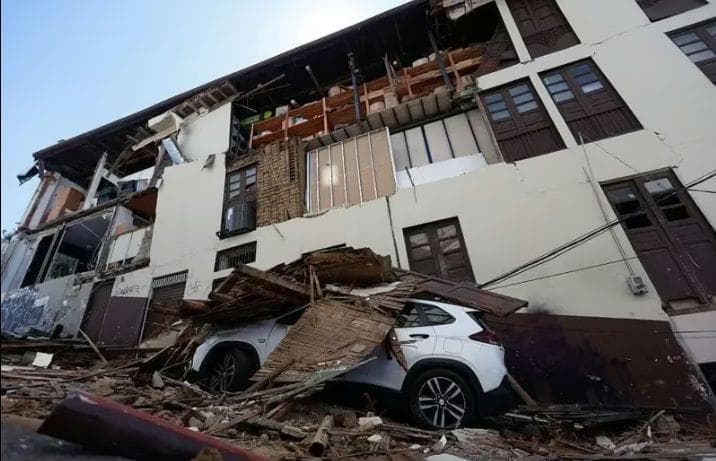 A residential block with collapsed upper floors, timber and masonry debris covering a parked car below.